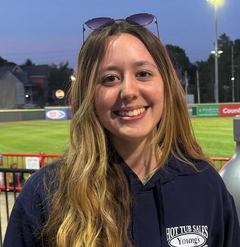 Smiling young woman in a navy hoodie with a Hot Tub Sales logo standing by a railing at a baseball stadium during dusk, with field and stadium lights in the background.