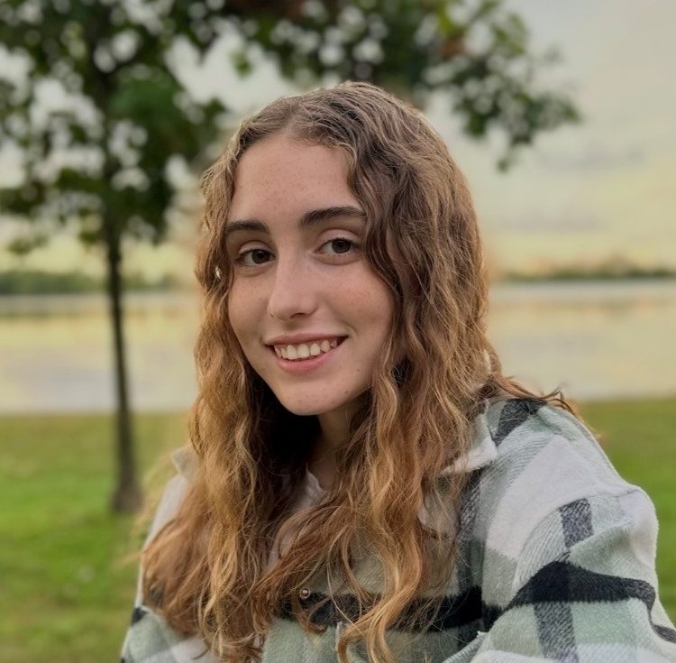Young woman with wavy hair in a plaid shirt sits on a wooden bench in a park by a lake, smiling at the camera.
