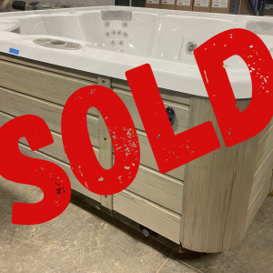 White hot tub on a wooden display in a warehouse, with a large red SOLD stamp across it indicating it’s sold.