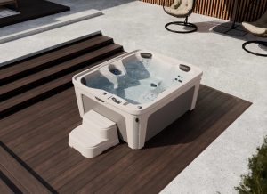 Outdoor beige hot tub on a wooden deck with step-stairs and nearby patio chairs in a backyard setting.