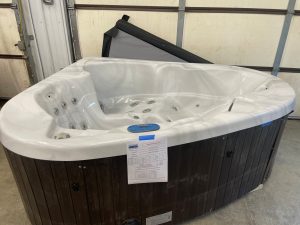 White hot tub in a showroom with a folded dark cover leaning against a wall and papers taped to the front panel.