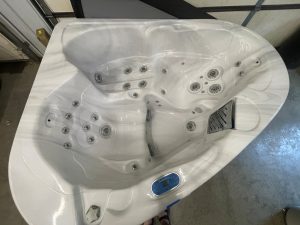 White hot tub with numerous circular water jets and built-in seating, featuring a blue digital control panel on the edge, indoors in a workshop/garage setting